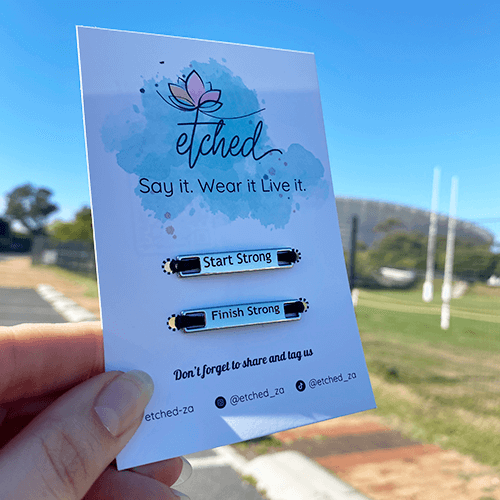 Image of etched product card showing the Start Strong Finish Strong shoe tags being held up by a hand with the Cape Town Stadium in the background